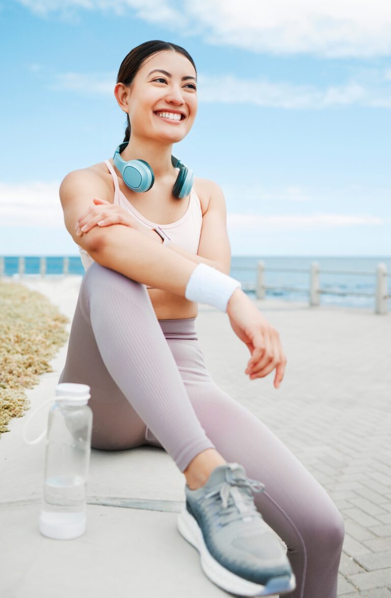 Fitness woman, happy and beach exercise while taking break or rest while out running on promenade w